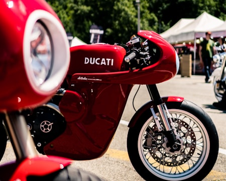 A striking red Ducati motorcycle is prominently displayed, showcasing its sleek design and engineering. The background features a blurred outdoor setting with tents and people, suggesting a bike show or event atmosphere.