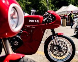 A striking red Ducati motorcycle is prominently displayed, showcasing its sleek design and engineering. The background features a blurred outdoor setting with tents and people, suggesting a bike show or event atmosphere.