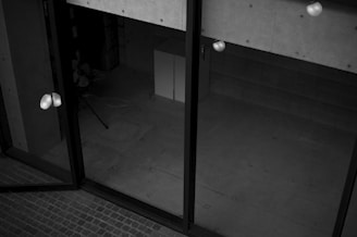 A black and white image depicting a modern architectural space with large glass doors. The interior appears empty, with visible concrete walls and a tiled floor. There are some light reflections on the glass surface and a few indistinct objects like a tripod or stand.