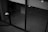 A black and white image depicting a modern architectural space with large glass doors. The interior appears empty, with visible concrete walls and a tiled floor. There are some light reflections on the glass surface and a few indistinct objects like a tripod or stand.