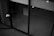 A black and white image depicting a modern architectural space with large glass doors. The interior appears empty, with visible concrete walls and a tiled floor. There are some light reflections on the glass surface and a few indistinct objects like a tripod or stand.