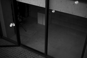 A black and white image depicting a modern architectural space with large glass doors. The interior appears empty, with visible concrete walls and a tiled floor. There are some light reflections on the glass surface and a few indistinct objects like a tripod or stand.