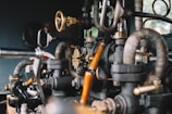 A close-up view of intricate metal pipes, valves, and gauges interconnected in a mechanical system. The image highlights the complexity and craftsmanship of the industrial components, showcasing a mix of brass and black metallic colors with a slightly rustic texture.