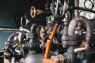 A close-up view of intricate metal pipes, valves, and gauges interconnected in a mechanical system. The image highlights the complexity and craftsmanship of the industrial components, showcasing a mix of brass and black metallic colors with a slightly rustic texture.