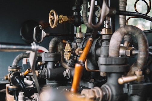 A close-up view of intricate metal pipes, valves, and gauges interconnected in a mechanical system. The image highlights the complexity and craftsmanship of the industrial components, showcasing a mix of brass and black metallic colors with a slightly rustic texture.