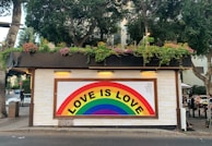 A small building with white brick walls adorned with a colorful rainbow mural featuring the text 'LOVE IS LOVE'. The roof has an abundance of greenery and flowers. There are trees surrounding the building, and some people are sitting and standing nearby. One person is seen walking on the sidewalk.