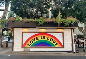 A small building with white brick walls adorned with a colorful rainbow mural featuring the text 'LOVE IS LOVE'. The roof has an abundance of greenery and flowers. There are trees surrounding the building, and some people are sitting and standing nearby. One person is seen walking on the sidewalk.