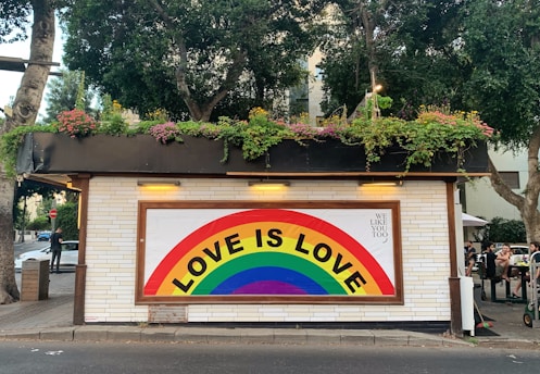 A small building with white brick walls adorned with a colorful rainbow mural featuring the text 'LOVE IS LOVE'. The roof has an abundance of greenery and flowers. There are trees surrounding the building, and some people are sitting and standing nearby. One person is seen walking on the sidewalk.