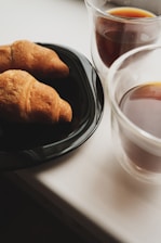 Two croissants resting on a black plate, accompanied by two cups of coffee in clear glass mugs. The setting appears to be on a light-colored tabletop, suggesting a cozy breakfast or snack environment.