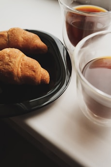 Two croissants resting on a black plate, accompanied by two cups of coffee in clear glass mugs. The setting appears to be on a light-colored tabletop, suggesting a cozy breakfast or snack environment.