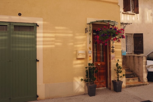 A charming facade of a house with a light yellow wall. A wooden door with intricate ironwork is surrounded by potted plants and a hanging flower basket bursting with pink and purple blooms. To the left is a green garage door, and a mailbox is attached to the wall beside the entrance. Sunlight casts soft shadows across the scene.