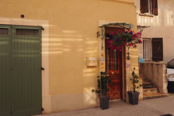A charming facade of a house with a light yellow wall. A wooden door with intricate ironwork is surrounded by potted plants and a hanging flower basket bursting with pink and purple blooms. To the left is a green garage door, and a mailbox is attached to the wall beside the entrance. Sunlight casts soft shadows across the scene.