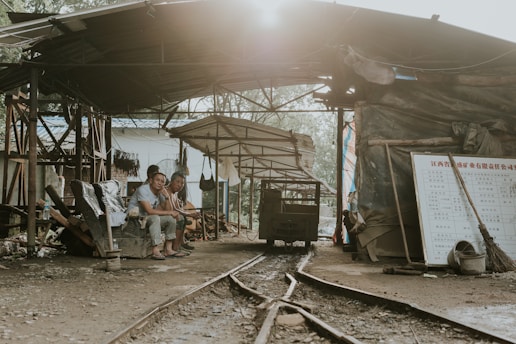 Three people are sitting under a makeshift shelter with corrugated metal roofing. The area appears to be part of an industrial or construction site, evidenced by the tracks running through and the various tools and materials scattered around. Sunlight filters through the roof, casting a warm glow.
