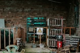 An industrial setting with exposed electrical equipment housed against a brick wall. The equipment consists of various transformers, circuit breakers, and wiring, enclosed within metal racks. A warning sign in a triangle is prominently displayed, indicating high voltage, and is written in Chinese. The scene has an abandoned and slightly rustic feel, with some rust and overgrown plants visible.