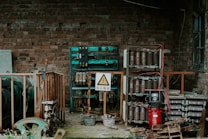 An industrial setting with exposed electrical equipment housed against a brick wall. The equipment consists of various transformers, circuit breakers, and wiring, enclosed within metal racks. A warning sign in a triangle is prominently displayed, indicating high voltage, and is written in Chinese. The scene has an abandoned and slightly rustic feel, with some rust and overgrown plants visible.