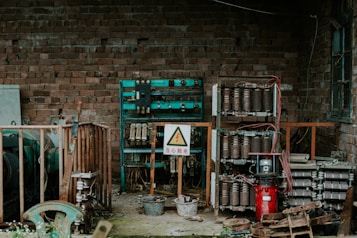 An industrial setting with exposed electrical equipment housed against a brick wall. The equipment consists of various transformers, circuit breakers, and wiring, enclosed within metal racks. A warning sign in a triangle is prominently displayed, indicating high voltage, and is written in Chinese. The scene has an abandoned and slightly rustic feel, with some rust and overgrown plants visible.