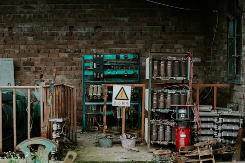 An industrial setting with exposed electrical equipment housed against a brick wall. The equipment consists of various transformers, circuit breakers, and wiring, enclosed within metal racks. A warning sign in a triangle is prominently displayed, indicating high voltage, and is written in Chinese. The scene has an abandoned and slightly rustic feel, with some rust and overgrown plants visible.