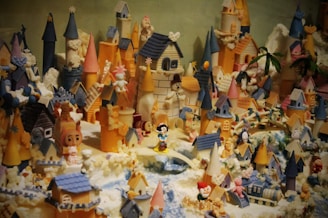 A whimsical and detailed miniature village filled with colorful castles, towers, houses, and various small figurines. The structures are decorated with an array of vibrant colors, including blue, pink, and orange. Numerous toy-like characters and animals are scattered throughout the scene, creating a playful and enchanted atmosphere.