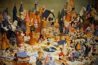 A whimsical and detailed miniature village filled with colorful castles, towers, houses, and various small figurines. The structures are decorated with an array of vibrant colors, including blue, pink, and orange. Numerous toy-like characters and animals are scattered throughout the scene, creating a playful and enchanted atmosphere.