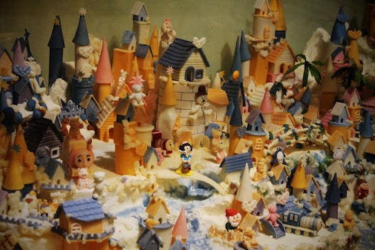 A whimsical and detailed miniature village filled with colorful castles, towers, houses, and various small figurines. The structures are decorated with an array of vibrant colors, including blue, pink, and orange. Numerous toy-like characters and animals are scattered throughout the scene, creating a playful and enchanted atmosphere.