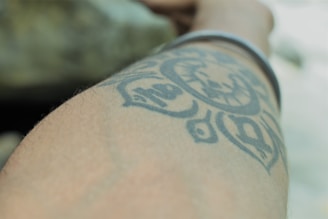 A close-up view of a forearm with a black tattoo featuring intricate patterns and designs. The tattoo appears to have circular elements and abstract shapes. A bracelet is visible on the wrist, and the background is blurred, focusing attention on the arm.