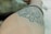 A close-up view of a forearm with a black tattoo featuring intricate patterns and designs. The tattoo appears to have circular elements and abstract shapes. A bracelet is visible on the wrist, and the background is blurred, focusing attention on the arm.