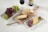 A variety of foods are artfully arranged on a white surface. A rectangular platter of red and green grapes is paired with a wooden board that holds a wedge of cheese and slices of baguette. Next to it, a marble tray displays slices of salami, cheese, and a small glass bowl of green olives. A glass of red wine and a bottle remain in the background, with two knives placed alongside the boards.