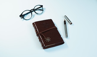A brown leather-bound journal with an embossed design and a decorative clasp is placed on a light background. Next to the journal, a pair of black-rimmed glasses and a silver pen with its cap off are neatly arranged.