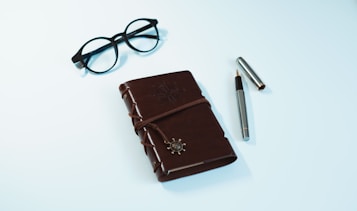A brown leather-bound journal with an embossed design and a decorative clasp is placed on a light background. Next to the journal, a pair of black-rimmed glasses and a silver pen with its cap off are neatly arranged.