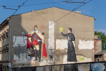 A street mural on a large, weathered wall depicts two historical figures. On the left, a regal man in a red cape and crown holds a selfie stick, creating a contrast between historical opulence and modern technology. On the right, a woman in traditional attire holds a glowing object resembling a light bulb, reflecting themes of innovation and enlightenment. The wall shows signs of decay, adding a vintage texture to the scene.