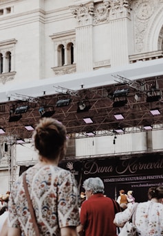 Several people stand in front of a stage with a large banner advertising the 'Duna Karneval' festival. The setting appears to be in front of an ornate historic building with intricate architectural details. There are stage lights visible above the people.