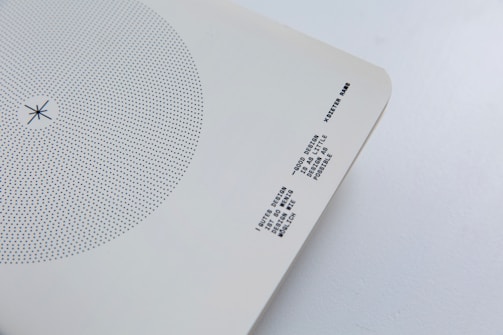 A minimalist design artifact featuring a circular pattern of blue dots on a white background. Near the bottom right corner, there is text printed in a monochrome typewriter-style font. The paper has slightly curved corners and is set against a light grey surface.