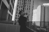 A person stands on an outdoor terrace, taking a photo with a camera. They are surrounded by several tall, modern skyscrapers. The architecture is sleek, with multiple windows and an urban cityscape setting. The image is in black and white, adding a classic and timeless feel.