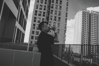 A person stands on an outdoor terrace, taking a photo with a camera. They are surrounded by several tall, modern skyscrapers. The architecture is sleek, with multiple windows and an urban cityscape setting. The image is in black and white, adding a classic and timeless feel.