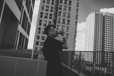 A person stands on an outdoor terrace, taking a photo with a camera. They are surrounded by several tall, modern skyscrapers. The architecture is sleek, with multiple windows and an urban cityscape setting. The image is in black and white, adding a classic and timeless feel.