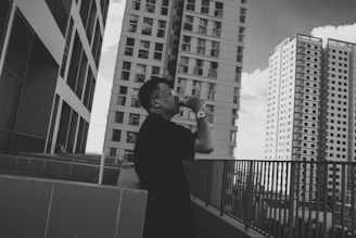 A person stands on an outdoor terrace, taking a photo with a camera. They are surrounded by several tall, modern skyscrapers. The architecture is sleek, with multiple windows and an urban cityscape setting. The image is in black and white, adding a classic and timeless feel.