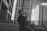 A person stands on an outdoor terrace, taking a photo with a camera. They are surrounded by several tall, modern skyscrapers. The architecture is sleek, with multiple windows and an urban cityscape setting. The image is in black and white, adding a classic and timeless feel.