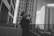 A person stands on an outdoor terrace, taking a photo with a camera. They are surrounded by several tall, modern skyscrapers. The architecture is sleek, with multiple windows and an urban cityscape setting. The image is in black and white, adding a classic and timeless feel.