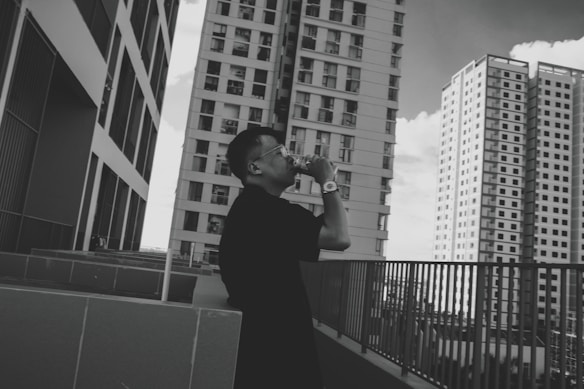 A person stands on an outdoor terrace, taking a photo with a camera. They are surrounded by several tall, modern skyscrapers. The architecture is sleek, with multiple windows and an urban cityscape setting. The image is in black and white, adding a classic and timeless feel.