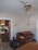A room with a piano against the wall beneath two wall lamps. A ceiling fan with gold and brass elements hangs from the ceiling. There is an open view into a kitchen, where a stove and some kitchen items are visible. A large black garbage bag sits on the floor near the piano.