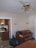 A room with a piano against the wall beneath two wall lamps. A ceiling fan with gold and brass elements hangs from the ceiling. There is an open view into a kitchen, where a stove and some kitchen items are visible. A large black garbage bag sits on the floor near the piano.