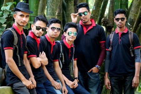 A group of six young men wearing matching black polo shirts with red collars and sunglasses, posing confidently next to a bamboo grove. Some carry backpacks while one wears a hat.