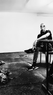 A person wearing a gas mask sits in a stark, minimally furnished room. The setting appears industrial or utilitarian, with a plain floor and wall. The person is wearing casual clothing, including checkered pants and sneakers, and casually leans forward with an arm resting on a piece of furniture. Various items lie scattered on the floor, contributing to a sense of disarray.
