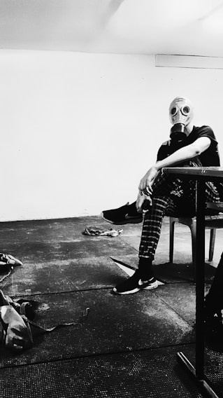 A person wearing a gas mask sits in a stark, minimally furnished room. The setting appears industrial or utilitarian, with a plain floor and wall. The person is wearing casual clothing, including checkered pants and sneakers, and casually leans forward with an arm resting on a piece of furniture. Various items lie scattered on the floor, contributing to a sense of disarray.