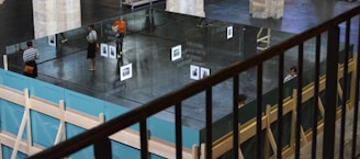 A modern art installation indoors featuring large mirrors forming a rectangular structure. Reflections of people and a black-and-white photo are visible on the mirrored surfaces. The surrounding space has columns and wooden support structures, with one person in an orange shirt standing nearby.