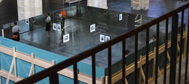 A modern art installation indoors featuring large mirrors forming a rectangular structure. Reflections of people and a black-and-white photo are visible on the mirrored surfaces. The surrounding space has columns and wooden support structures, with one person in an orange shirt standing nearby.