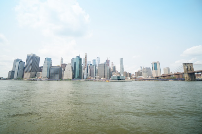 A panoramic view of a city skyline featuring modern skyscrapers and a suspension bridge. The buildings vary in height and architectural style, standing behind a wide body of water under a partly cloudy sky.