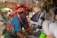 Women dressed in colorful traditional clothing and headscarves are washing dishes at an outdoor sink. They are surrounded by tents and a thatched-roof shelter, suggesting a campsite setting.