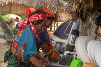 Women dressed in colorful traditional clothing and headscarves are washing dishes at an outdoor sink. They are surrounded by tents and a thatched-roof shelter, suggesting a campsite setting.