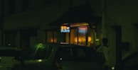 A dimly lit street scene featuring a sushi restaurant with a neon sign. Cars are parked outside, and a person stands near the entrance. Warm yellow and orange lights illuminate the interior from the windows.
